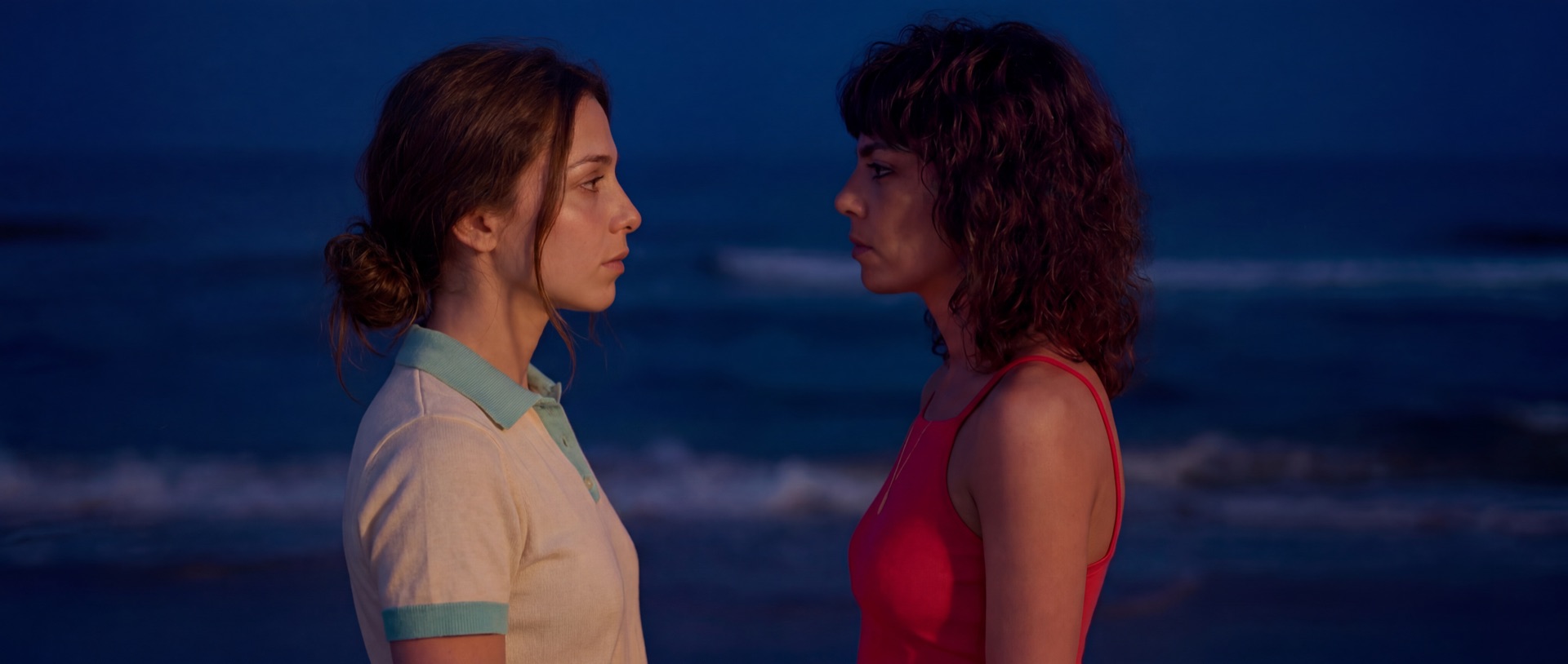 Two women face one another on a beach at blue hour